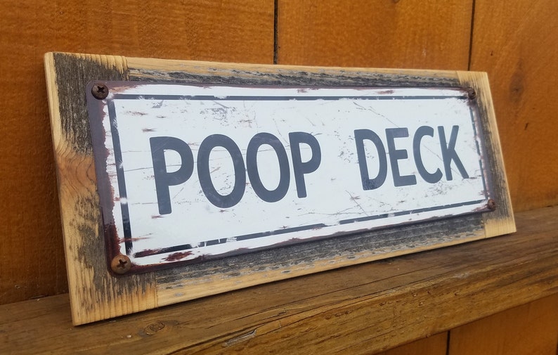 Puede incluir: Un letrero de madera r&uacute;stico con una placa de metal blanco que dice "POOP DECK". El letrero est&aacute; desgastado y tiene un aspecto envejecido.
