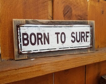May include: A rustic wooden sign with a white background and black text that reads "Born to Surf". The sign has a distressed look and is nailed to a wooden surface.