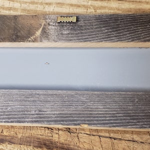 Puede incluir: La parte posterior de un marco rectangular de madera con un fondo gris. El marco tiene un aspecto r&uacute;stico con nudos y vetas visibles. Hay cuatro piezas triangulares de madera unidas a la parte posterior del marco para colgarlo.