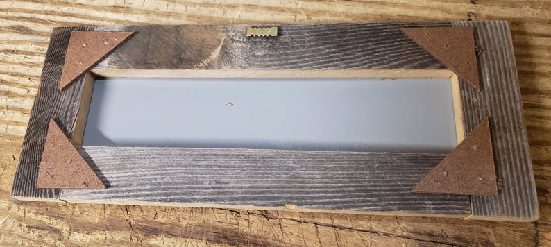 Puede incluir: La parte posterior de un marco rectangular de madera con un fondo gris. El marco tiene un aspecto r&uacute;stico con nudos y vetas visibles. Hay cuatro piezas triangulares de madera unidas a la parte posterior del marco para colgarlo.