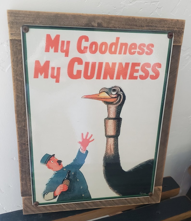 Puede incluir: Un letrero de metal vintage con un marco de madera marr&oacute;n. El letrero presenta una ilustraci&oacute;n de dibujos animados de una avestruz con un cuello largo y un hombre sosteniendo una botella de Guinness. El texto en el letrero dice "My Goodness My GUINNESS".