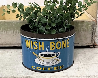 Op de afbeelding: Een blauwe metalen blik met een geel en wit ontwerp met een kopje koffie en de tekst "Wish Bone Coffee". Het blik is gevuld met groene bladplanten.