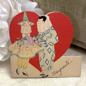 May include: Vintage Valentine's Day card featuring a heart-shaped design with two figures in costume. One figure wears a diamond-patterned outfit, and the other wears a yellow dress. The card is tan with the name "Marguerite" written in cursive.