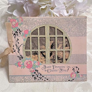 May include: A vintage-style greeting card with a pink background and floral accents. The card features a circular window design with a detailed interior scene. The text "Just Peepin' in to Cheer You!" is printed on the front.