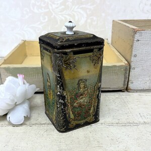 May include: Vintage tin box with a black and gold design. The box features a white knob on the lid and a colorful illustration of two girls in a field of flowers.