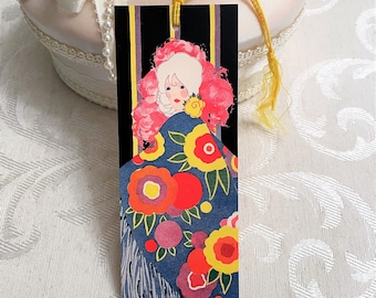 May include: A vintage bookmark featuring a woman with pink hair wearing a blue and red floral patterned shawl. The bookmark has black and gold stripes on the sides.