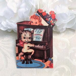 May include: Vintage Valentine's Day card featuring a cartoon cat playing a piano with the words "Be My Valentine" on the music sheet. The card is decorated with red and blue flowers.