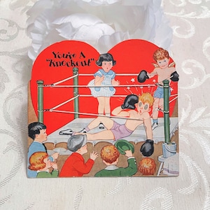 May include: A vintage Valentine's Day card featuring a red heart-shaped boxing ring with a group of children dressed as boxers. The text "You're A "Knockout" is printed on the card.