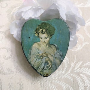 May include: A heart-shaped tin box with a vintage illustration of a woman with long hair, wearing a white dress, on a blue background. The tin box has a blue and green color scheme and the text "The Pierrot" is printed on the bottom.