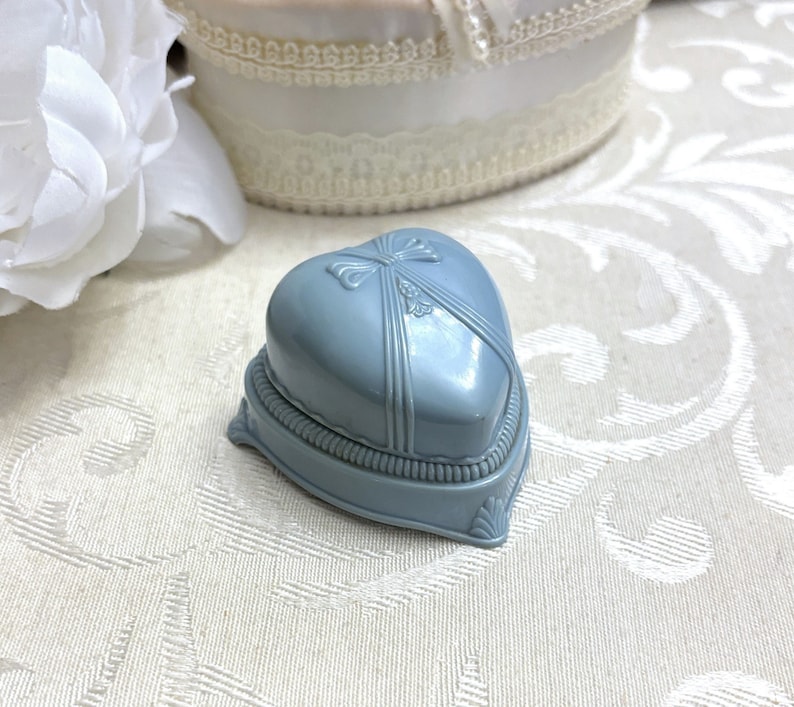 May include: A light blue heart-shaped ring box with a decorative bow on the lid. The box is made of plastic and has a satin-like finish.