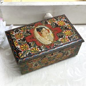 May include: A rectangular metal box featuring a colorful floral and geometric pattern in red, yellow, blue, and green. The lid displays a central oval portrait of a person wearing a lace veil and flowers in their hair, set against a red background.
