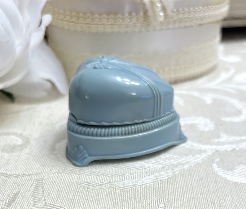 May include: A light blue plastic ring box with a heart-shaped lid. The box has a decorative trim around the edge and a small design on the front.