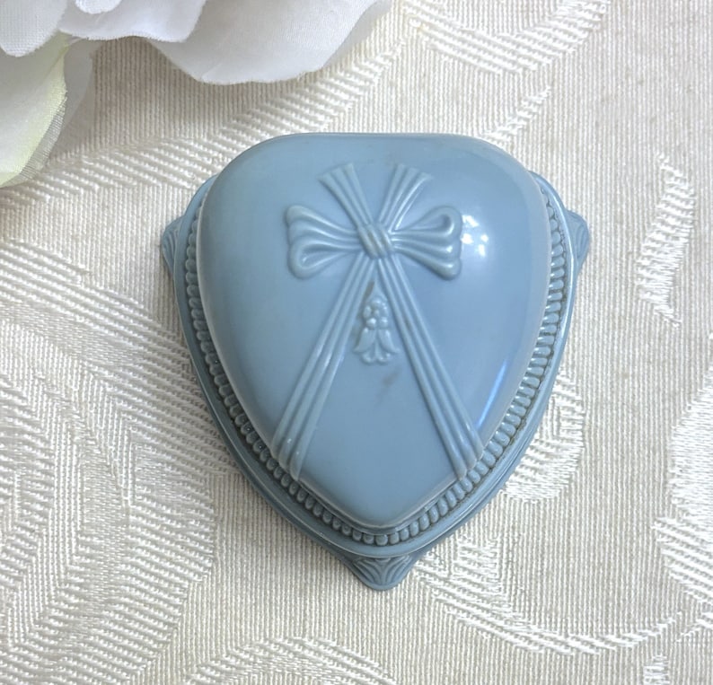 May include: A light blue heart-shaped ring box with a raised bow design and a beaded trim.
