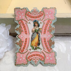 May include: A vintage paper cutout with a delicate, intricate design. The cutout features a woman in a blue dress and hat, holding a bird. The design is in shades of pink, gold, and white, with a blue background.