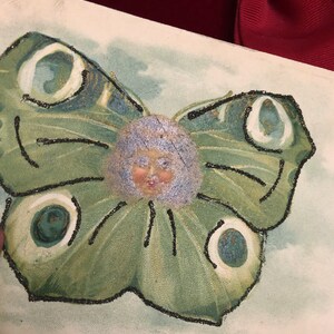Puede incluir: Ilustraci&oacute;n vintage de una mariposa con rostro humano. La mariposa tiene alas verdes con contornos de purpurina negra y detalles en blanco y verde azulado. La cara tiene cabello azul claro y una cara rosada.