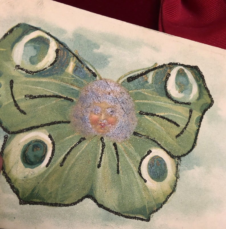 Puede incluir: Ilustraci&oacute;n vintage de una mariposa con rostro humano. La mariposa tiene alas verdes con contornos de purpurina negra y detalles en blanco y verde azulado. La cara tiene cabello azul claro y una cara rosada.