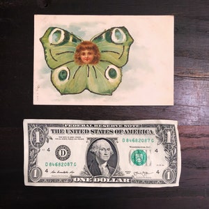 Puede incluir: Postal vintage con una ilustraci&oacute;n caprichosa de la cara de un ni&ntilde;o dentro de las alas de una mariposa. La mariposa es principalmente verde con detalles blancos y negros. Debajo hay un billete de un d&oacute;lar.