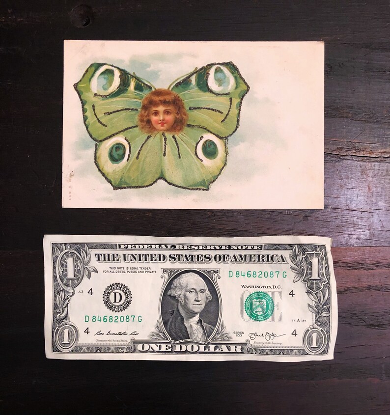 Puede incluir: Postal vintage con una ilustraci&oacute;n caprichosa de la cara de un ni&ntilde;o dentro de las alas de una mariposa. La mariposa es principalmente verde con detalles blancos y negros. Debajo hay un billete de un d&oacute;lar.