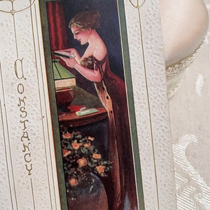 K&ouml;nnte beinhalten: Eine Vintage-Postkarte mit gr&uuml;nem Rand, die eine Frau in einem langen Kleid zeigt, die in einem Raum mit einer Lampe und Blumen steht. Der Text "Constancy" ist auf der linken Seite der Postkarte gedruckt.