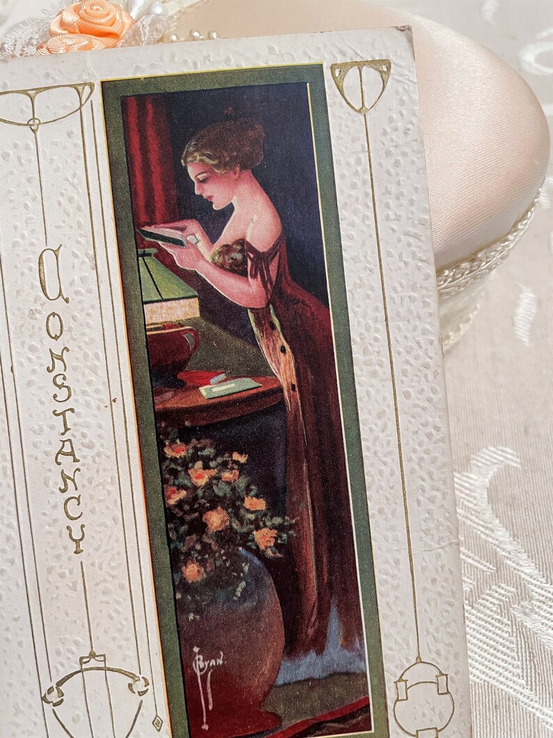 K&ouml;nnte beinhalten: Eine Vintage-Postkarte mit gr&uuml;nem Rand, die eine Frau in einem langen Kleid zeigt, die in einem Raum mit einer Lampe und Blumen steht. Der Text "Constancy" ist auf der linken Seite der Postkarte gedruckt.