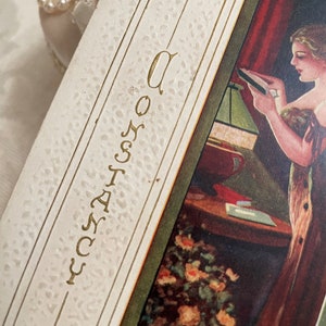 K&ouml;nnte beinhalten: Eine Vintage-Postkarte mit goldfarbenem Rand und dem Wort "Constancy" vertikal in Kursivschrift geschrieben. Die Postkarte zeigt eine Frau, die in einem schwach beleuchteten Raum einen Brief neben einer Lampe liest.