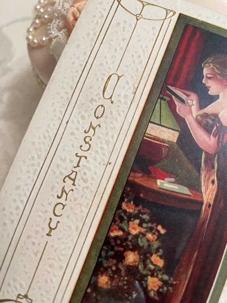 K&ouml;nnte beinhalten: Eine Vintage-Postkarte mit goldfarbenem Rand und dem Wort "Constancy" vertikal in Kursivschrift geschrieben. Die Postkarte zeigt eine Frau, die in einem schwach beleuchteten Raum einen Brief neben einer Lampe liest.
