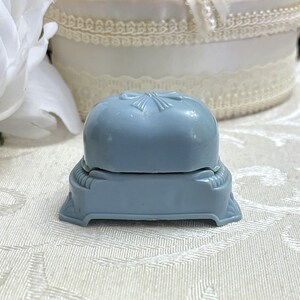 May include: A light blue ring box with a raised bow design on the lid. The box is made of plastic and has a hinged lid.
