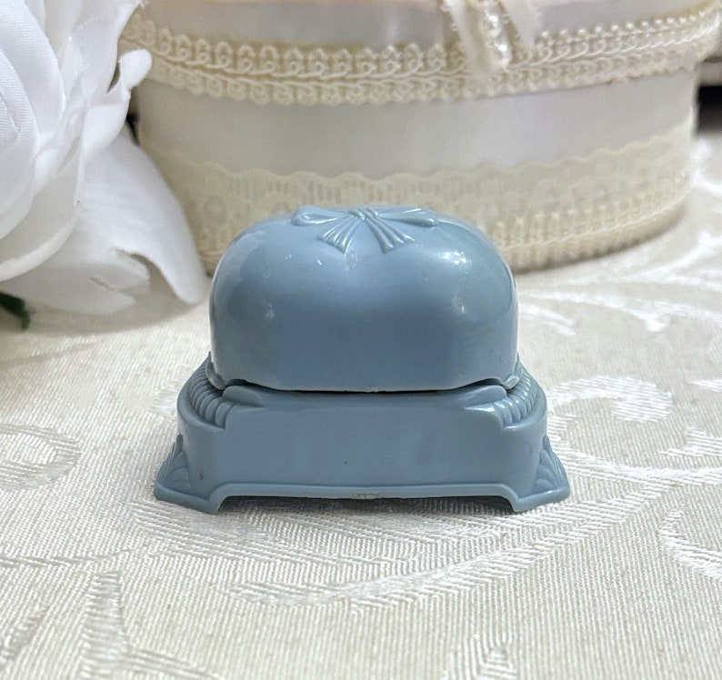 May include: A light blue ring box with a raised bow design on the lid. The box is made of plastic and has a hinged lid.