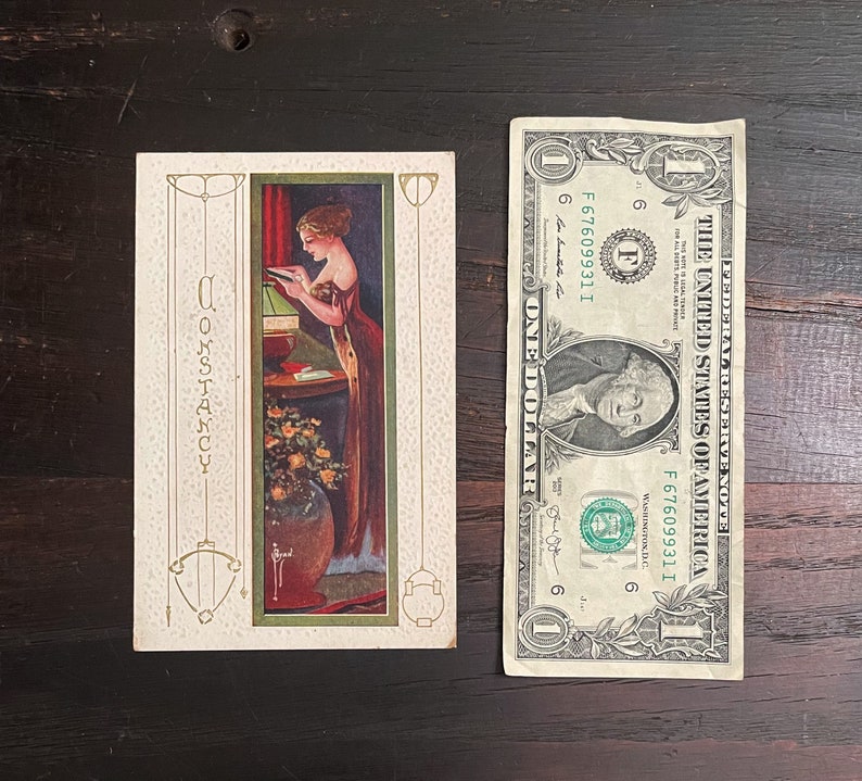 K&ouml;nnte beinhalten: Eine Vintage-Postkarte mit einer Frau in einem langen Kleid, die ein Buch liest. Die Postkarte hat einen goldenen Rand und das Wort "Constancy" steht vertikal auf der linken Seite.