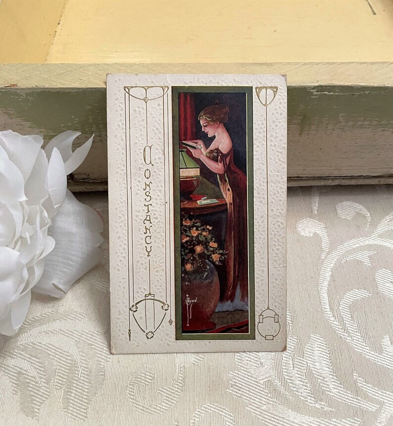 K&ouml;nnte beinhalten: Vintage-Postkarte mit einer Frau in einem langen roten Kleid, die an einem Tisch mit einer Blumenvase einen Brief liest. Die Postkarte hat das Wort "Constancy" vertikal auf der linken Seite geschrieben.