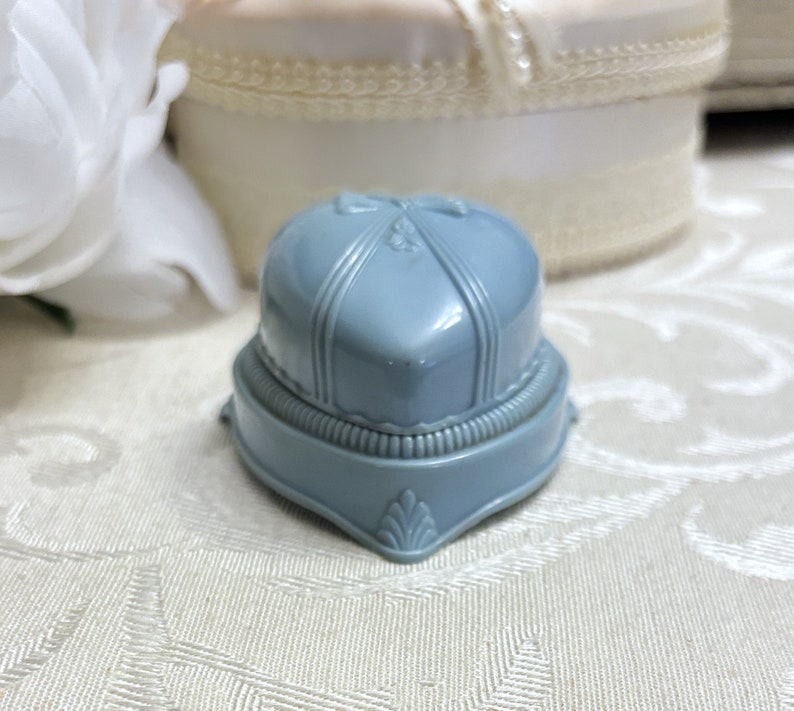 May include: A light blue ring box with a heart-shaped lid and decorative details. The box is made of plastic and has a vintage look.