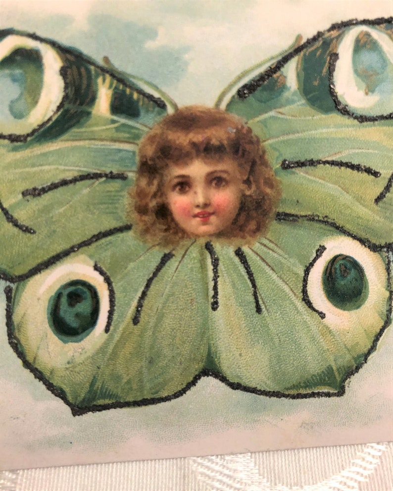 Puede incluir: Ilustraci&oacute;n vintage de la cara de un ni&ntilde;o rodeada de alas de mariposa. Las alas son principalmente verdes con detalles de purpurina negra. El ni&ntilde;o tiene cabello casta&ntilde;o rizado y mejillas rosadas, sobre un fondo azul claro.