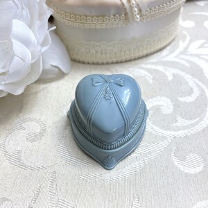 May include: A light blue heart-shaped ring box with a satin ribbon and bow on the lid. The box has a decorative design around the edge.
