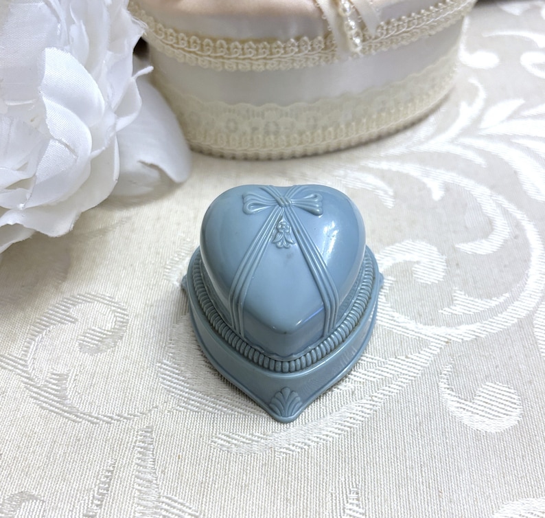 May include: A light blue heart-shaped ring box with a satin ribbon and bow on the lid. The box has a decorative design around the edge.
