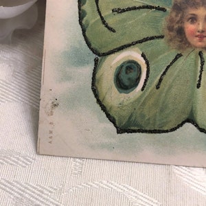 Puede incluir: Postal vintage con la cara de una ni&ntilde;a rodeada por un dise&ntilde;o de ala de mariposa verde. Las alas de la mariposa tienen detalles de purpurina negra. El fondo es azul claro y blanco.