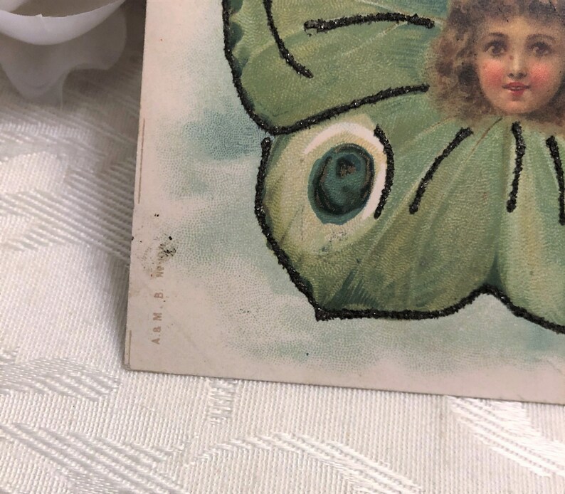 Puede incluir: Postal vintage con la cara de una ni&ntilde;a rodeada por un dise&ntilde;o de ala de mariposa verde. Las alas de la mariposa tienen detalles de purpurina negra. El fondo es azul claro y blanco.