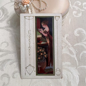 K&ouml;nnte beinhalten: Vintage-Postkarte mit einer Frau, die ein Buch neben einer Lampe liest. Die Postkarte hat einen gr&uuml;nen Rand und das Wort "Constancy" steht vertikal auf der linken Seite.