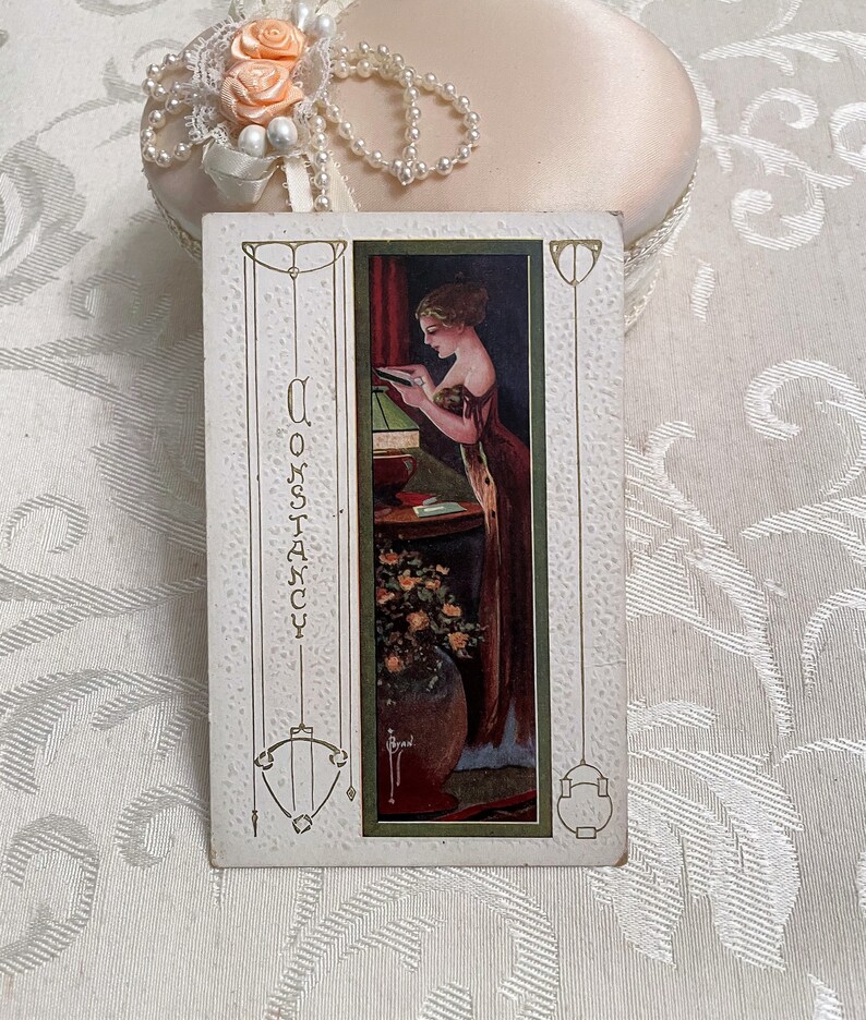 K&ouml;nnte beinhalten: Vintage-Postkarte mit einer Frau, die ein Buch neben einer Lampe liest. Die Postkarte hat einen gr&uuml;nen Rand und das Wort "Constancy" steht vertikal auf der linken Seite.