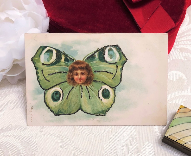 Puede incluir: Postal vintage que representa una mariposa verde con la cara de un ni&ntilde;o en el centro. Las alas de la mariposa est&aacute;n detalladas con contornos verde oscuro y patrones circulares. El ni&ntilde;o tiene cabello casta&ntilde;o rizado y una expresi&oacute;n suave. La postal tiene un fondo claro.