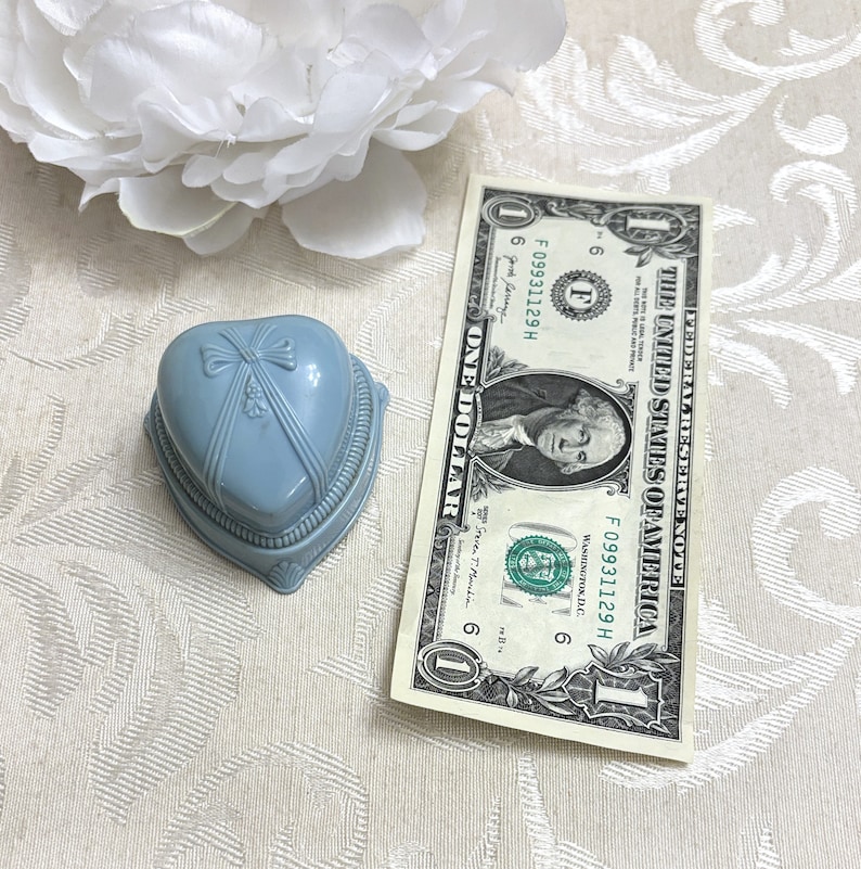 May include: A light blue heart-shaped ring box with a white bow on the lid. The box is sitting on a white surface with a one dollar bill next to it.