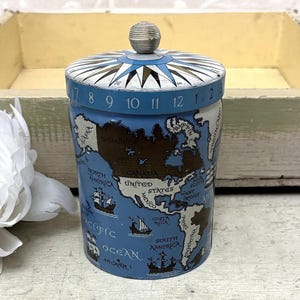 May include: A cylindrical metal tin featuring a vintage-style map of North and South America in shades of blue and brown. The lid displays a compass rose design and a small silver knob handle. The rim of the lid is printed with numbers 1 through 12.