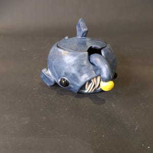 May include: A whimsical, blue ceramic sugar bowl shaped like a fish. The bowl features a lid with a fin-like handle, black eyes, and a yellow ball. The mouth reveals white teeth. The sugar bowl is a unique kitchen decor item.