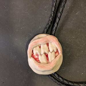 May include: A ceramic bolo tie featuring a grinning mouth with white teeth and pink lips. The mouth is attached to a black braided leather cord. The teeth are set against a red background.