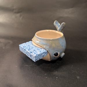 May include: A ceramic whale-shaped sponge holder with a glossy blue and cream glaze. The whale features a raised tail fin, a dark eye, and an open mouth designed to hold a rectangular blue kitchen sponge, which is currently inserted inside.