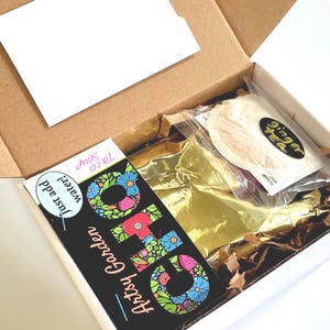May include: An open cardboard box containing a package of ginger tea, a card with the text "Artsy Garden" and "Just add water!" and a packet of taco soup. The box is lined with gold foil and brown packing material.