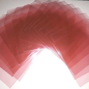 May include: A fan of translucent, reddish-pink plastic sheets, fanned out against a white background. The sheets are square or rectangular, with a slight sheen, and appear to be stacked. The light catches the edges, creating highlights and shadows.