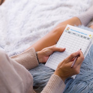 Puede incluir: Una persona sentada en una cama con una manta blanca y peluda, escribiendo en una hoja de papel blanca con un bolígrafo negro. El papel tiene un patrón de cuadrícula y el texto "Secure Your Deal Wish List" en la parte superior.
