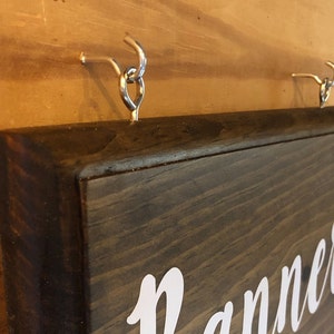 May include: A dark brown wooden sign with the word "Planner" written in white cursive lettering. The sign is hanging from two silver hooks.