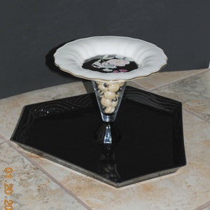 May include: A white ceramic serving dish with a floral design sits on top of a black octagonal tray. The dish is filled with small, round, beige objects.