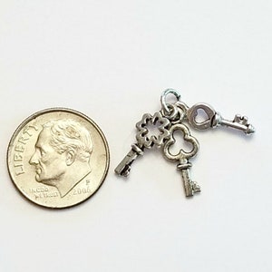 May include: Three silver key charms with ornate designs, including a heart-shaped keyhole, a clover, and a traditional key. The charms are shown next to a US dime for scale.